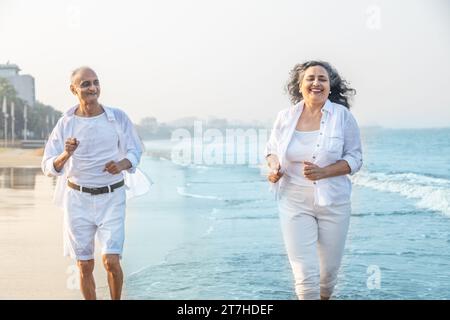 Glückliches, älteres indisches Paar, das weiße Tücher trägt, läuft zusammen am Strand und hat Spaß. Urlaub und Gesundheitskonzept Stockfoto