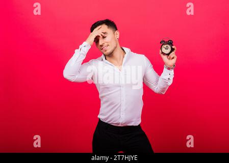 Emotionaler junger Mann, der die Uhr auf rotem Hintergrund hält. Das Konzept der Verspätung Stockfoto