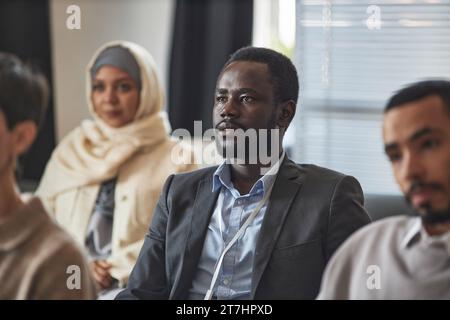 Junger afroamerikanischer männlicher Angestellter in Formalwear, der dem Sprecher zuhört, während er zwischen multiethnischen männlichen Kollegen und muslimischen Frauen sitzt Stockfoto