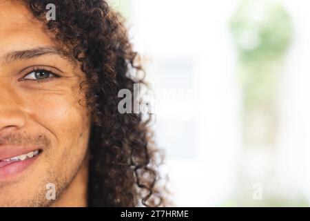 Halbgesicht eines glücklichen birassischen Mannes mit langen dunklen lockigen Haaren lächelnd im sonnigen Wohnzimmer, Kopierraum Stockfoto