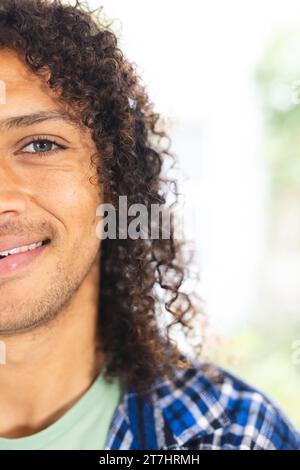 Halbgesicht eines glücklichen birassischen Mannes mit langen dunklen lockigen Haaren lächelnd im sonnigen Wohnzimmer, Kopierraum Stockfoto