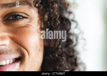 Halbgesicht eines glücklichen birassischen Mannes mit langen dunklen lockigen Haaren lächelnd im sonnigen Wohnzimmer, Kopierraum Stockfoto