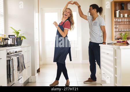Glückliches, abwechslungsreiches Paar, das in der sonnigen Küche zu Hause tanzt und lächelt Stockfoto