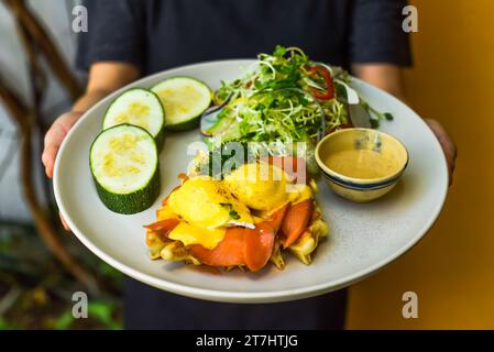 Kellnerin serviert im Restaurant einen Teller mit Eiern, bennedict-Waffeln und geräuchertem Lachs Stockfoto