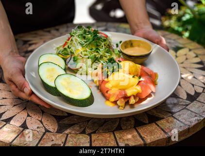 Kellnerin serviert im Restaurant einen Teller mit Eiern, bennedict-Waffeln und geräuchertem Lachs Stockfoto