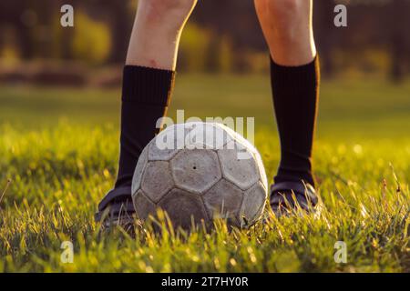 Unerkennbare junge Athletin in hohen Socken steht am frühen Sommermorgen vor dem Fußball auf gemähtem Gras. Sport und Gesundheit. Kinderbereich Stockfoto