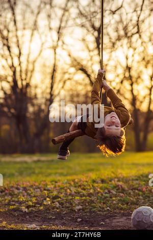 Ein lächelnder Teenager hängt kopfüber auf einer hausgemachten Schaukel in der Natur, im Park, im Dorf bei Sonnenuntergang. Spiele und Unterhaltung im Freien. Glücklich Stockfoto