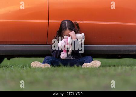 Das kleine einsame Mädchen weint, versteckt ihr Gesicht in Spielzeug, sitzt auf Gras und drückt den Rücken gegen das Auto. Kindertränen. Das Kind ist verärgert und beleidigt Stockfoto