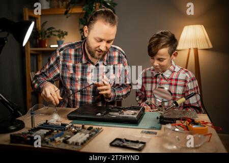 Der glückliche Junge und sein Vater reparieren die Ausstellungskarte mit einem Lötkolben, der zusammen am Tisch sitzt. Stockfoto