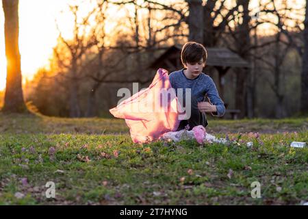 Freiwilligenarbeit für Kinder. Der Teenager sammelt Müll in einem großen rosafarbenen Beutel in der Natur, auf der Wiese am Herbstabend bei Sonnenuntergang. Umweltprobleme Stockfoto