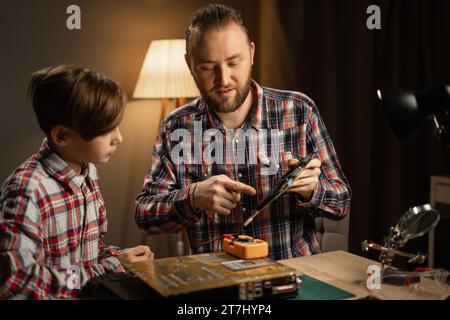 Der kaukasische Vater und sein Sohn verwenden Lötkolben, um das Motherboard am Laptop zu befestigen. Familie sitzt zusammen am Tisch und repariert den Computer zu Hause. Stockfoto