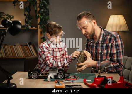 Vater und Sohn in Schutzbrille mit Lötkolben zur Reparatur von Spielzeugauto zu Hause am Abend. Familienbegriff und Zweisamkeit. Stockfoto