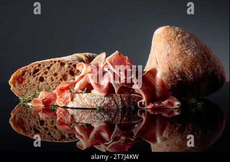 Prosciutto mit Ciabatta und Thymian auf schwarzem reflektierenden Hintergrund. Stockfoto