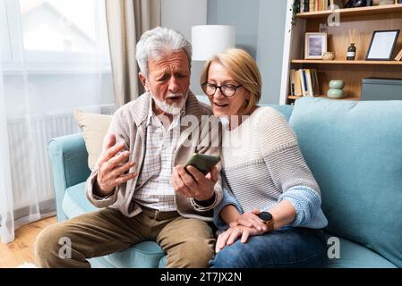 Glückliches älteres Ehepaar, das Smartphone hält und auf den Handy-Bildschirm schaut lachend entspannt sitzen zusammen auf dem Sofa sitzen, lächeln ältere Großeltern Familie Stockfoto