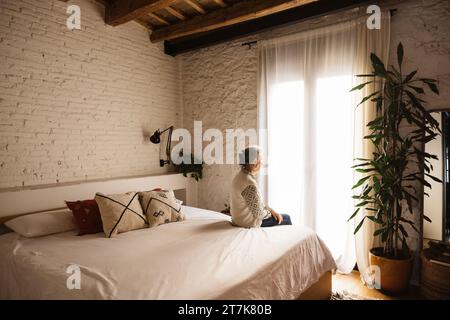 Porträt einer nachdenklichen, echten älteren Frau, die auf dem Bett vor einem Fenster in einem Schlafzimmer sitzt. Stockfoto