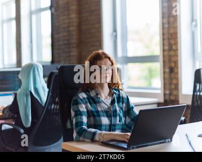 Eine junge und erfolgreiche Geschäftsfrau mit leuchtend orangefarbenen Haaren arbeitet in einem modernen Büro fokussiert und zeigt ihr Selbstvertrauen und ihre Dynamik Stockfoto