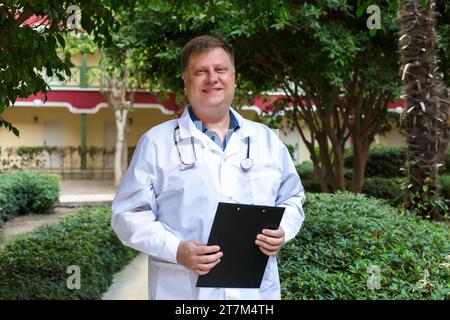 Ein dicker blonder männlicher Arzt mit Dokumenten auf dem Krankenhaushof. Stockfoto