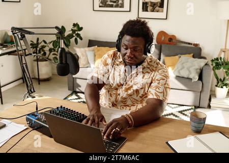 Seriöser afroamerikanischer Typ mit Kopfhörern, der auf der Laptop-Tastatur sitzt, während er am Arbeitsplatz im Studio oder zu Hause sitzt und Podcasts aufnimmt Stockfoto