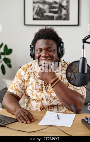Junger, lächelnder afroamerikanischer Moderator oder Radiomoderator in Kopfhörern, der am Schreibtisch sitzt und im Mikrofon mit dem Gast spricht Stockfoto