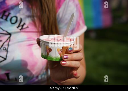 Nahaufnahme einer Frauenhand, die eine Tasse rosa Eiscreme hält. Die Hand verfügt über leuchtende, funkelnde Nägel in den Farben Rot, Orange, Violett und Blau Stockfoto