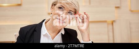 Glücklicher blonder Immobilienmakler mittleren Alters mit Brille, der in die Kamera im Immobilienbüro schaut, Banner Stockfoto