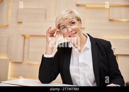 Glücklicher blonder Immobilienmakler mittleren Alters mit Brillen, der in einem Immobilienbüro arbeitet und in die Kamera schaut Stockfoto