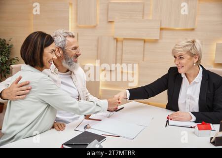 Glückliches verheiratetes Paar, das dem Makler die Hand schüttelt und im Immobilienbüro einen Handel macht, Haus kauft Stockfoto