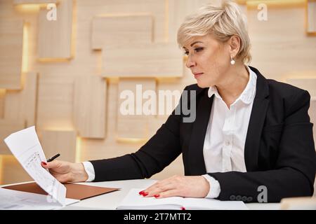 Blonder Makler mittleren Alters mit kurzen Haaren arbeitet in einem Immobilienbüro und sieht sich den Plan an Stockfoto