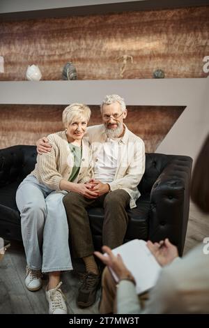 Glückliches verheiratetes Paar mittleren Alters, das sich während der Familientherapie umarmt und auf einer Ledercouch sitzt Stockfoto