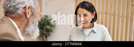 Glückliche Frau mittleren Alters lächelt in der Nähe des bärtigen Maklers in der Immobilienagentur, Banner Stockfoto