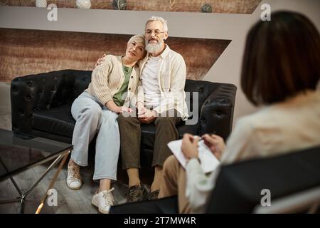 Glückliches Paar mittleren Alters, das sich während der Familientherapie umarmt und auf einer Ledercouch sitzt Stockfoto