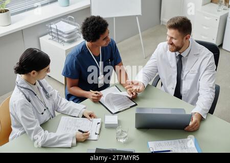 Hochwinkelansicht bei drei professionellen jungen Ärzten, die sich am Tisch in der Klinik treffen und den Patientenfall diskutieren Stockfoto