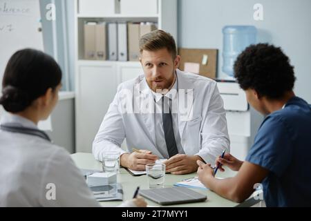 Porträt eines bärtigen männlichen Arztes, der einen Labormantel trägt und junge Auszubildende beim Treffen anweist Stockfoto
