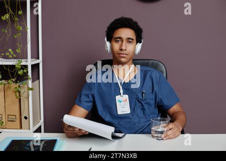 Vorderansicht des jungen Mannes aus dem Nahen Osten als männlicher Arzt, der am Schreibtisch im Büro sitzt und Kopfhörer mit Geräuschminimierung trägt Stockfoto