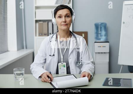 Porträt einer reiferen Ärztin, die mit dem Patienten per Videochat am Arbeitsplatz spricht und Kopfhörer trägt, Kopierraum Stockfoto