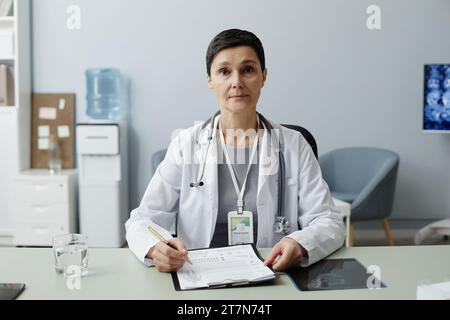 Frontporträt der Reifen Frau als Ärztin, die am Arbeitsplatz in der Klinik sitzt und in die Kamera blickt Stockfoto