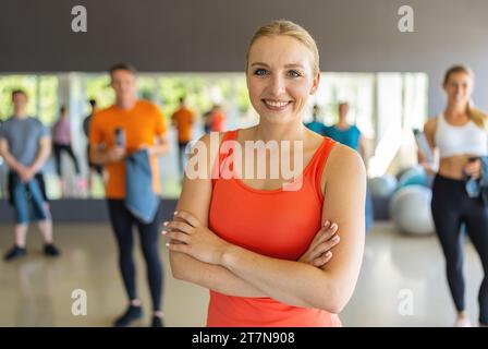 Selbstbewusste Frau in orangefarbenem Tanktop, stehend mit überkreuzten Armen in einem Fitnessstudio. Teamwork-Konzept-Bild Stockfoto