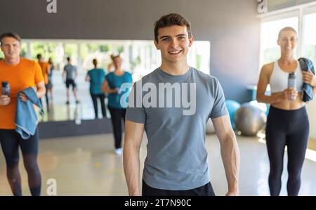 Junger Mann in grauem T-Shirt lächelt in einem Fitnessstudio mit anderen Leuten im Hintergrund. Teamwork-Konzept-Bild Stockfoto
