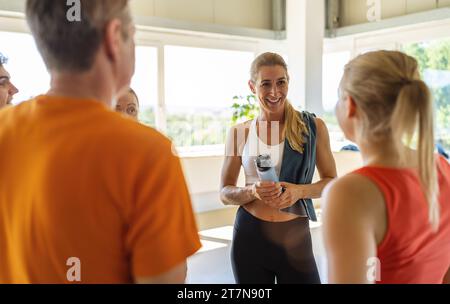 Lächelnde Frau mit einer Wasserflasche, die mit anderen Fitnessmitgliedern in einem sonnigen Raum spricht Stockfoto