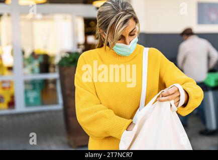 Frau, die ihre Stofftasche überprüft, während sie eine Maske vor einem Laden trägt Stockfoto