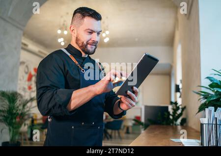 Lächelnder Inhaber eines kleinen Unternehmens in schwarzer Chefuniform mit Schürze und digitalem Tablet in seinem gemütlichen Restaurantsaal. Erfolgreiche Menschen, hart W Stockfoto