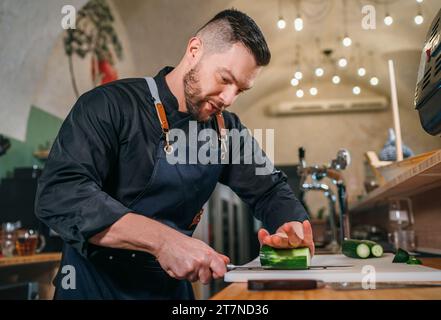 Bärtiger Koch in schwarzer Uniform mit einer Schürze, die frische grüne Gurke mit scharfem Messer in der Küche des Restaurants im Keller serviert. Professionell Stockfoto