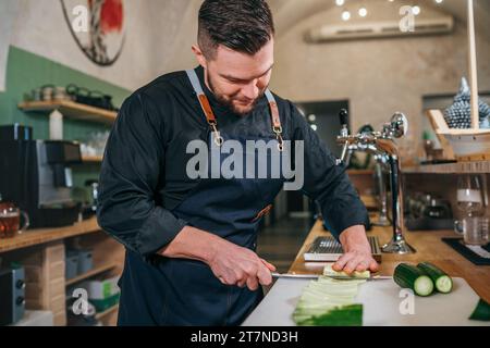 Bärtiger Koch in schwarzer Uniform mit einer Schürze, die frische grüne Gurke mit scharfem Messer in der Küche des Restaurants im Keller serviert. Professionell Stockfoto