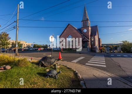 Zwei bronzefarbene Schildkrötenplastiken, eine große und eine kleine, stehen auf einem Verkehrsinsel gegenüber der in einen Veranstaltungsort umgewandelten Backsteinkirche in Marquette, Michigan, Vereinigte Staaten. Eine orangefarbene Verkehrszapfenkappe sitzt oben auf dem Stoppschildpfosten hinter ihnen. Stockfoto