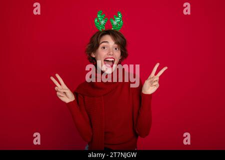 Schönes junges Mädchen in Weihnachten Rentier Hörner und Schal posiert und genießt auf Party auf rotem Hintergrund. Frohe weihnachten. Frohes neues Jahr. Urlaub Stockfoto