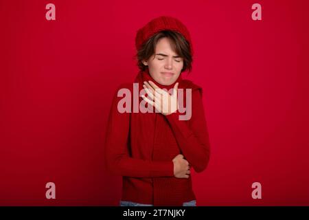 Nahaufnahme junge traurige Frau trägt roten Pullover, Schal und Hut bedeckt Mund mit Händen niedlicher Husten isoliert auf rotem Hintergrund im Studio. Gesundheit, krank, krank, Stockfoto