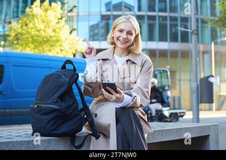 Porträt einer jungen glücklichen Studentin, Frau sitzt auf der Bank mit Rucksack und Telefon, zeigt mit dem Finger auf die linke obere Ecke, zeigt Werbung oder Stockfoto