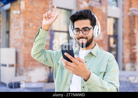 Ein junger Mann mit Kopfhörern läuft fröhlich durch die Stadt, lächelt, hört online Musik, verwendet eine Anwendung am Telefon. Stockfoto