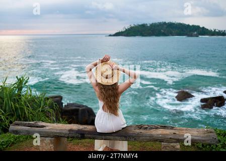 Frau in weißem Kleid und Strohhut sitzt auf einer Holzbank am Meer und genießt die Aussicht bei Sonnenuntergang. Weibliche Reisende entspannen sich an der tropischen Küste, Ruhe Stockfoto