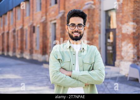 Porträt eines jungen Mannes in lässiger Kleidung, fröhlicher Geschäftsmann, der lächelnd und mit gekreuzten Armen in die Kamera blickt, indischer Mann vor dem Bürogebäude. Stockfoto